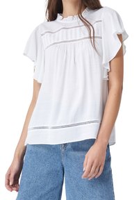 Blouse blanche avec des manches à volants, découpes horizontales et un col froncé. Tissu texturé avec un léger éclat, associé à un jean taille haute.
