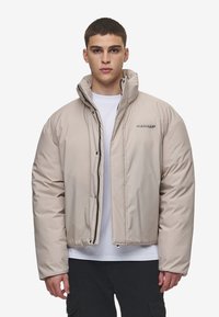 Jachetă puffer izolată bej, cu guler înalt, fermoar frontal și accent cu logo. Are o textură netedă și lucioasă, precum și o croială lejeră.