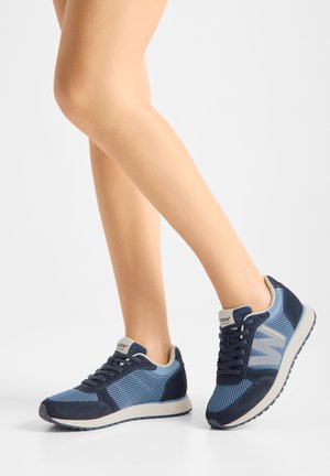 Gambe con sneakers in mesh blu e blu navy con suole bianche, posate con un piede piatto e l'altro leggermente sollevato su uno sfondo bianco.