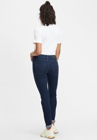Jeans de mezclilla azul oscuro con una cinturilla elástica, diseño ajustado y dos bolsillos traseros. Combinados con una camiseta blanca de manga corta y puños doblados.