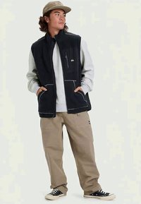 Melnā tekstūrētā veste pāri gaiši pelēkam džemperim, beiges bikses un melnas sporta apavi. Iezīmēta ar ar iezipjamajām kabatām, sānu kabatām un kontrastējošu šuvumu.