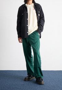 Person wearing black buttoned jacket, cream Adidas t-shirt, green track pants with white stripes, and black platform shoes standing on blue carpet.