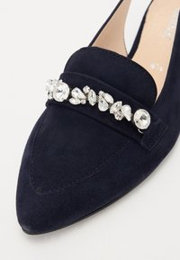 Mocasín de ante azul marino con punta afilada, que presenta un adorno decorativo de strass en la parte superior. Suela plana con costuras sutiles.