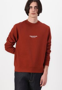 Rustkleurige sweatshirt van zacht materiaal, met een ronde hals, lange mouwen en witte "Originals Studio" branding op de borst.