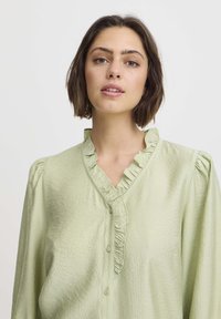 Light green blouse with ruffled V-neck, button front, and puffed sleeves. Textured fabric adds subtle detail.