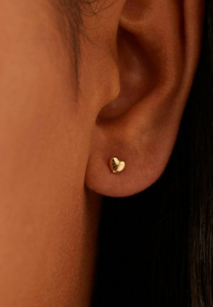 Close-up van een gouden oorbel in de vorm van een hartje, gedragen op de oorschelp tegen een achtergrond van mediumbruine huid en donker haar.