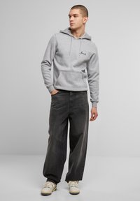Felpa grigia con cappuccio, cordino e tasca frontale, abbinata a jeans neri larghi e sneakers bianche, con un semplice logo sul petto.