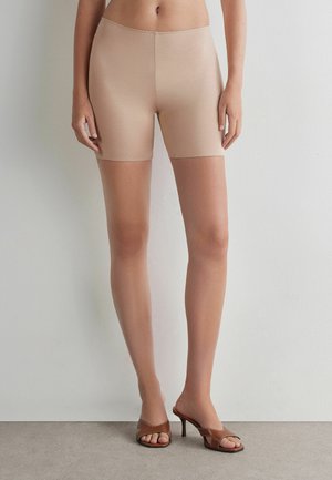 Kvinde iført beige tætsiddende shorts og brune høje hælede sandaler, stående op ad en hvid væg og grå gulv.