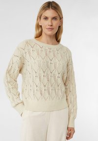 Cremfarbener gestrickter Pullover mit einem offenen Rautenmuster, geripptem Saum und Bündchen sowie einem runden Ausschnitt, kombiniert mit hellen Hosen.