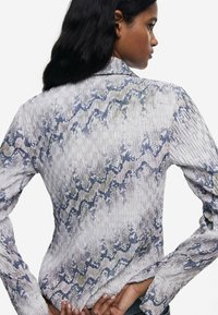 Ľahká, vzorovaná blúzka s textúrovaným povrchom v odtieňoch modrej, bielej a metalických akcentoch. Dlhé rukávy a golier.