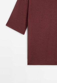 Gros plan sur le côté gauche d'un t-shirt uni à manches courtes en tissu rouge foncé sur fond blanc.