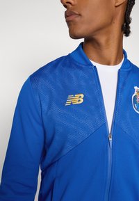 Blå treningjakke med glidelås, featuring pustende meshpaneler, gulllogo på brystet og avrundet krage. Glatt stoff med sporty design.
