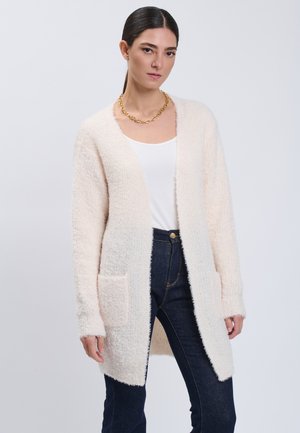 Woman wearing a light pink fuzzy long cardigan over white top and dark jeans, with a gold chain necklace and hair tied back.
