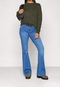 Maglione lavorato a maglia verde oliva con onde testurizzate, abbinato a jeans svasati blu e scarpe platform nere. Inclusi cintura nera e borsa a tracolla.