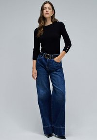 Haut noir à manches longues avec accents de boutons, associé à un jean en denim à jambes larges de couleur bleu foncé. Ceinture noire, talons noirs brillants. Design simple et contemporain.