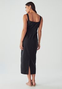 Ženska z dolgimi temnimi lasmi, ki nosi zlate kroglice in brezrokavno črno midi obleko z razporkom na hrbtu ter ravne sandale, stoji bočno.