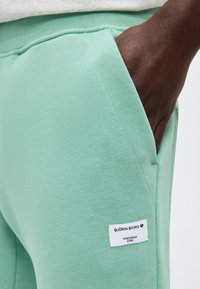 Χέρι με σκοτεινό δέρμα στην τσέπη ανοιχτοπράσινου παντελονιού φόρμας Björn Borg με λευκή ετικέτα της μάρκας στον μηρό.