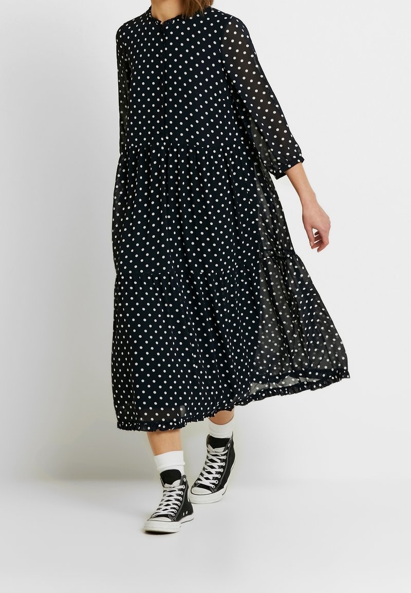 Femme marchant dans une robe midi noire à pois blancs, manches trois-quarts, chaussettes blanches et baskets montantes noires.