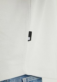 Biała siateczkowa koszula z teksturowanym wzorem, widocznymi szwami oraz czarną metką z małym białym logo po boku. W zestawie z niebieskimi dżinsami.