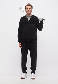 Jeune homme en veste et pantalon noirs tenant un club de golf sur l'épaule, portant un gant blanc et des baskets blanches, debout devant un fond blanc.