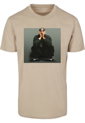 Beige bomulds t-shirt med et fremtrædende grafisk print af en siddende person iført sort tøj, hænderne på ansigtet, med sølvsmykker og tilbehør.