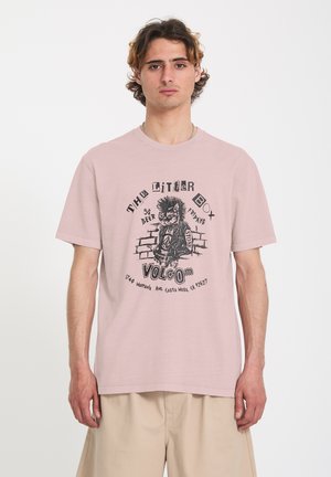 Jeune homme portant un t-shirt rose clair avec un dessin de bande dessinée punk noir et un texte sur un bar à l'avant, associé à un short beige.