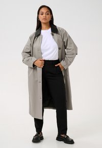 Grå trenchcoat med svart krage, med stora knappar och en avslappnad passform. Bärs över en vit T-shirt och svarta byxor, med kraftiga svarta skor.