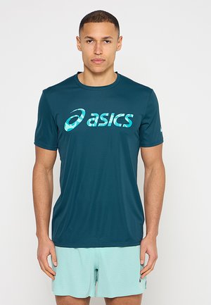 Uomo che indossa una maglietta sportiva Asics verde acqua e pantaloncini atletici azzurri chiaro, in piedi su uno sfondo bianco neutro.