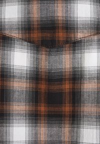 Tejido de cuadro que presenta colores negro, blanco y naranja con patrones de espiga. La textura parece suave y con relieve, adecuada para ropa o accesorios.