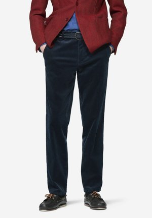 Man wearing dark blue corduroy pants, black belt, black boat shoes, red blazer, and blue shirt, standing with hands in pockets.