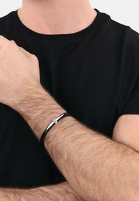 Pulsera de cuero negro con un acento plateado horizontal, llevada en una muñeca con cabello castaño claro. Diseño simple y moderno.