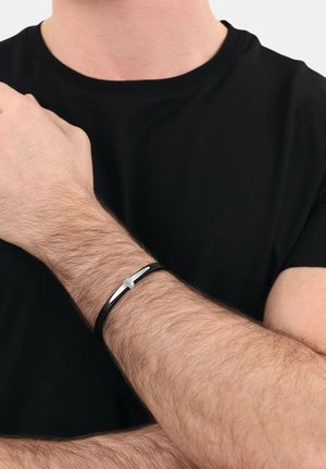 Pulsera de cuero negro con un acento plateado horizontal, llevada en una muñeca con cabello castaño claro. Diseño simple y moderno.