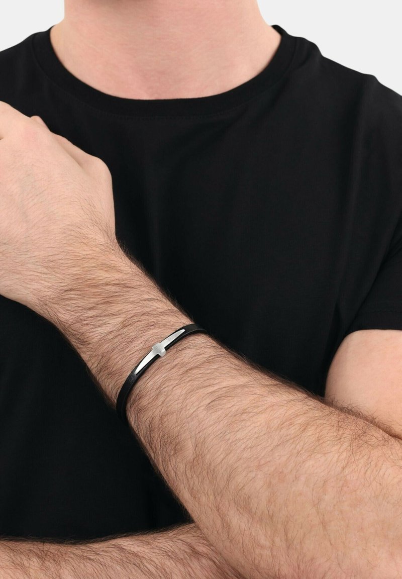 Pulsera de cuero negro con un acento plateado horizontal, llevada en una muñeca con cabello castaño claro. Diseño simple y moderno.