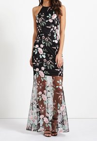 Robe maxi noire à fleurs avec un superposé transparent et brodé présentant des fleurs roses et vertes. Design sans manches et silhouette longue et fluide.