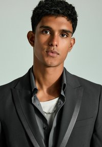 Chaqueta de traje gris con solapas de satén, superpuesta a una camisa gris de botones. Camiseta de cuello redondo texturizada blanca visible debajo, con detalle de cadena plateada.