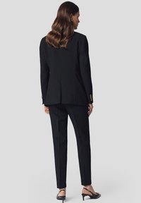 Czarny, dopasowany garnitur z usztywnioną marynarką z rozcięciem i dopasowanymi, wąskimi spodniami. W komplecie czarne buty na obcasie.