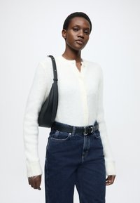 Κρεμ ζακέτα με ανάλαφρη υφή και κουμπιά μπροστά, συνδυασμένη με σκούρο μπλε τζιν ψηλής μέσης και κομψή μαύρη τσάντα ώμου.