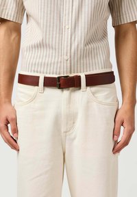 Brązowy skórzany pasek z metalową klamrą, noszony na jasnych, teksturowanych spodniach oraz na koszulce w paski z krótkim rękawem.