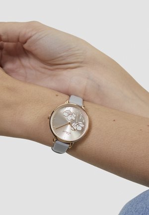 Round watch with a rose gold case and a light gray leather strap. The face features floral details and metallic hour markers.