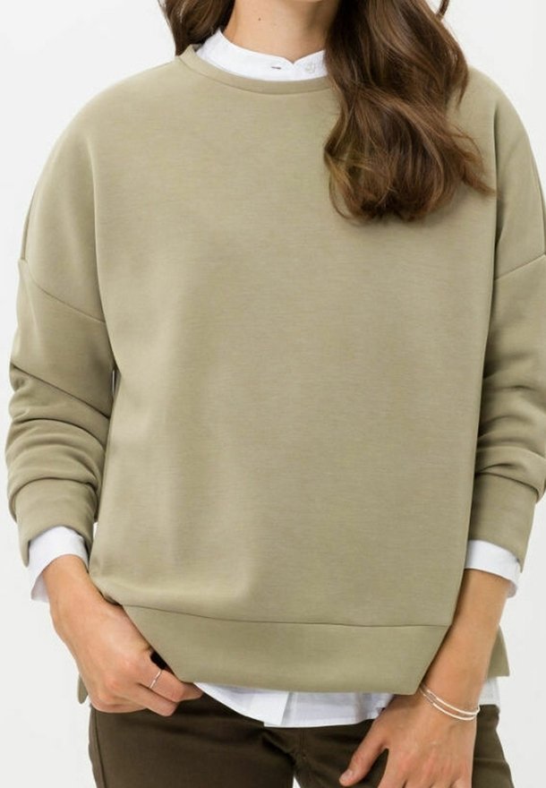 Femme portant un sweat-shirt vert olive clair sur une chemise blanche à col, avec un pantalon marron et peu de bijoux aux mains.