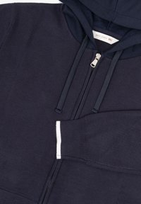 Marineblå zip-up hættetrøje med en stor hætte, ribbede manchetter og frontlomme. Har en sølvfarvet lynlås og kontrastende hvid accent på lommen.
