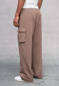 Baggy cargobroek in een lichtbruine stof met twee zijzakken, een elastische tailleband en witte sneakers. Getextureerd detail bij de zakranden.