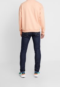 Person med ljusrosa sweatshirt, mörkblå tajta jeans och vita sneakers med gröna och orangea accenter, stående mot vit bakgrund.