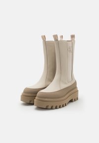 Beige ankelstøvler med elastiske sidepaneler, en teksturert brun såle og et rent, strømlinjeformet design, med en subtil logo detalj.