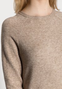 Beige strikkegenser med rund hals, ribbestrikkede mansjetter og subtil tekstur. Har raglanermer og en myk avslutning.