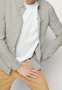 Ljusgrå linneskjorta med knäppning framtill, parat med en vit t-shirt med rund halsringning. Beiga chinos fulländar outfiten. Inga anmärkningsvärda mönster eller detaljer.