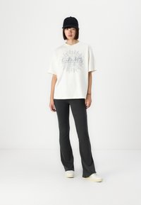 Overstørrelse creme t-shirt med sølv grafisk design, parret med tætsiddende charcoalsgrå flared bukser og hvide sneakers. En sort kasket fuldender looket.