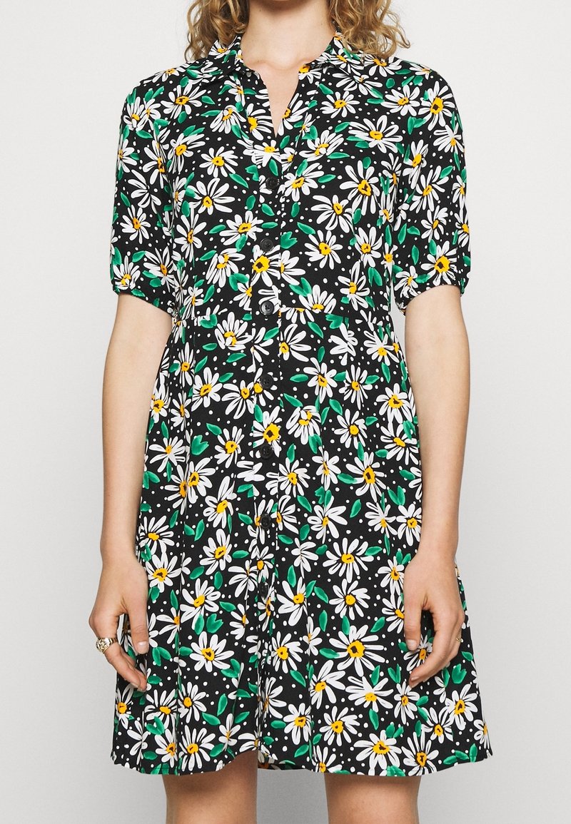 Femme portant une robe noire boutonnée avec un imprimé marguerite blanc et jaune et des manches courtes bouffantes, debout devant un fond uni.