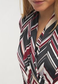 Barevný geometrický top ve odstínech burgundské, černé, krémové a bílé; má zavinovací střih s diagonálními pruhy a jemnou texturu látky.