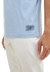 T-shirt en coton bleu clair à manches courtes, avec une étiquette imprimée en bas à gauche. Assorti avec un pantalon blanc.